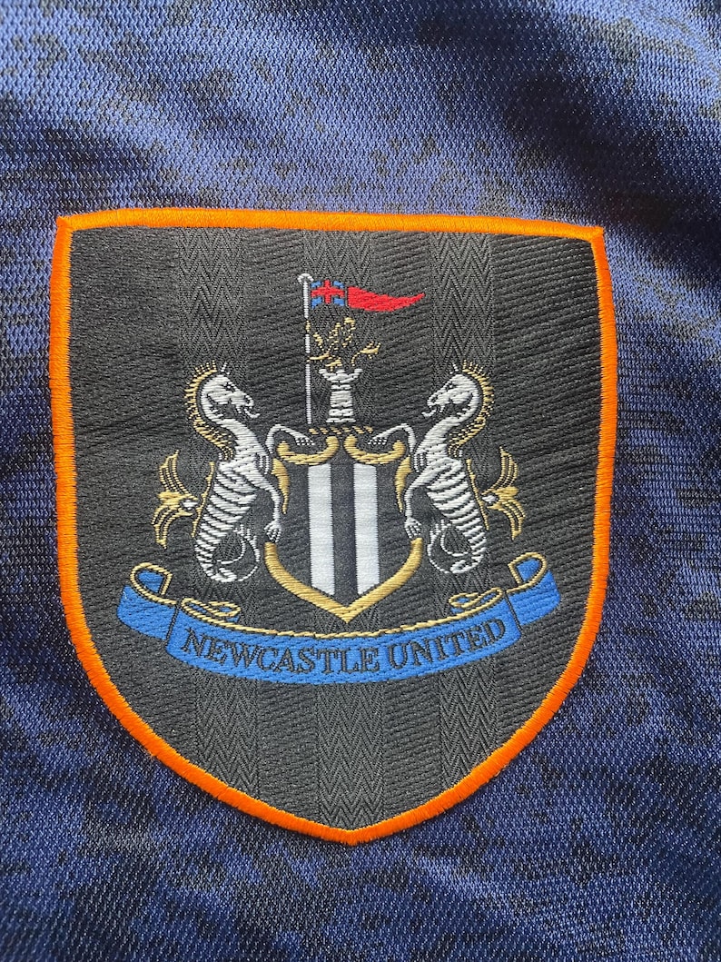 May include: Embroidered patch featuring the Newcastle United crest. The crest is shield-shaped with a black and white striped center, flanked by two seahorses. The patch has an orange border and a blue banner with the text "NEWCASTLE UNITED".