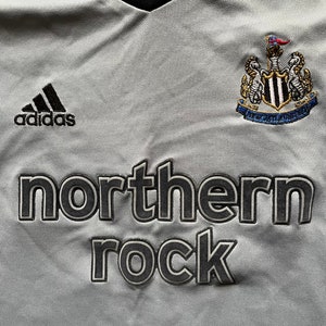 Pu&ograve; includere: Una maglia da calcio grigia con logo Adidas nero, uno stemma bianco e nero con una bandiera e il testo "northern rock" in nero.