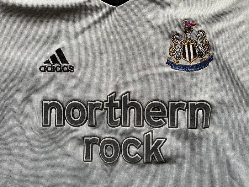 Pu&ograve; includere: Una maglia da calcio grigia con logo Adidas nero, uno stemma bianco e nero con una bandiera e il testo "northern rock" in nero.