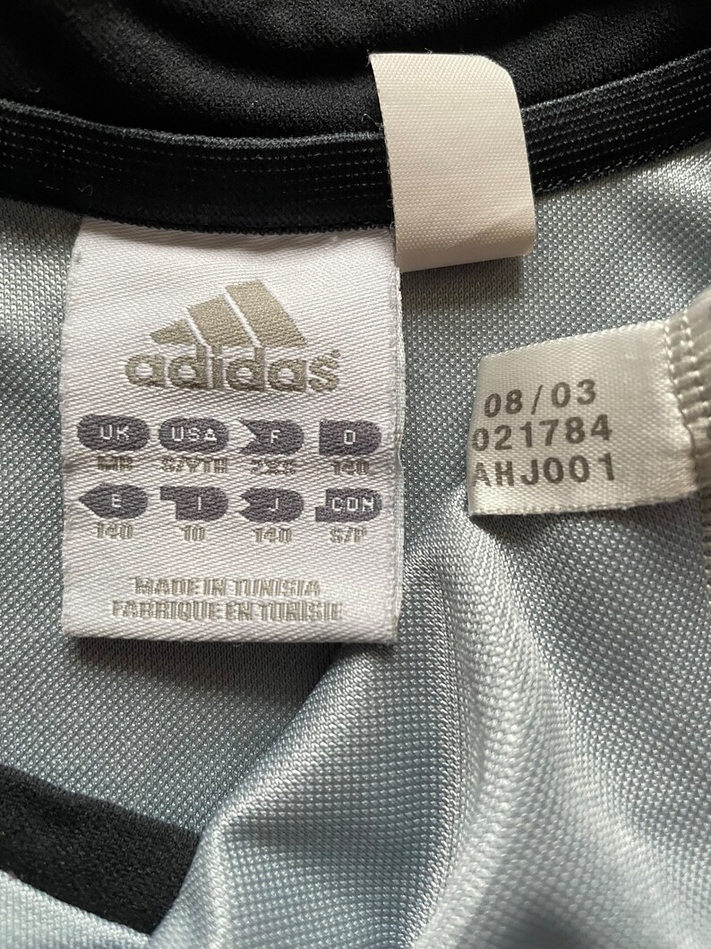 Pu&ograve; includere: Etichetta bianca con logo adidas nero e testo. L'etichetta contiene informazioni sulle taglie in taglie UK, USA, F, D, E, I, J e CON. L'etichetta contiene anche il testo "Made in Tunisia, Fabrique en Tunisie" e la data 08/03, 021784, AHJ001.