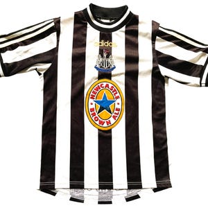 May include: A black and white striped soccer jersey with the Adidas logo and a Newcastle Brown Ale emblem. The jersey has a round neck and short sleeves with black stripes. The Newcastle Brown Ale emblem is a gold circle with a blue star.