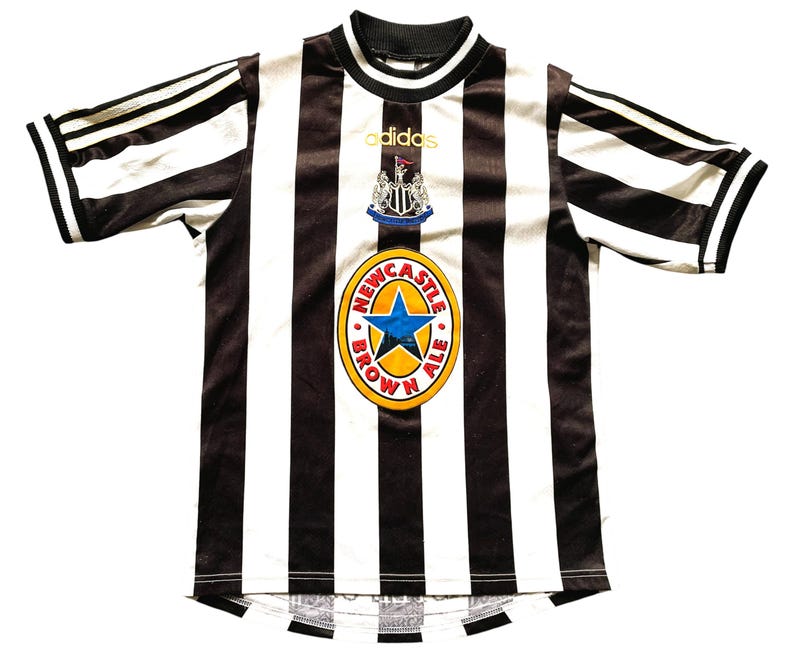 May include: A black and white striped soccer jersey with the Adidas logo and a Newcastle Brown Ale emblem. The jersey has a round neck and short sleeves with black stripes. The Newcastle Brown Ale emblem is a gold circle with a blue star.