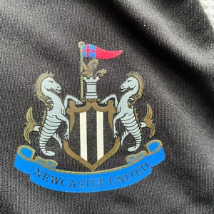 Pu&ograve; includere: Primo piano di un tessuto nero con lo stemma del Newcastle United. Lo stemma presenta due cavallucci marini, uno scudo a strisce bianche e nere e uno stendardo blu con la scritta "Newcastle United". Una bandiera rossa e blu in alto.