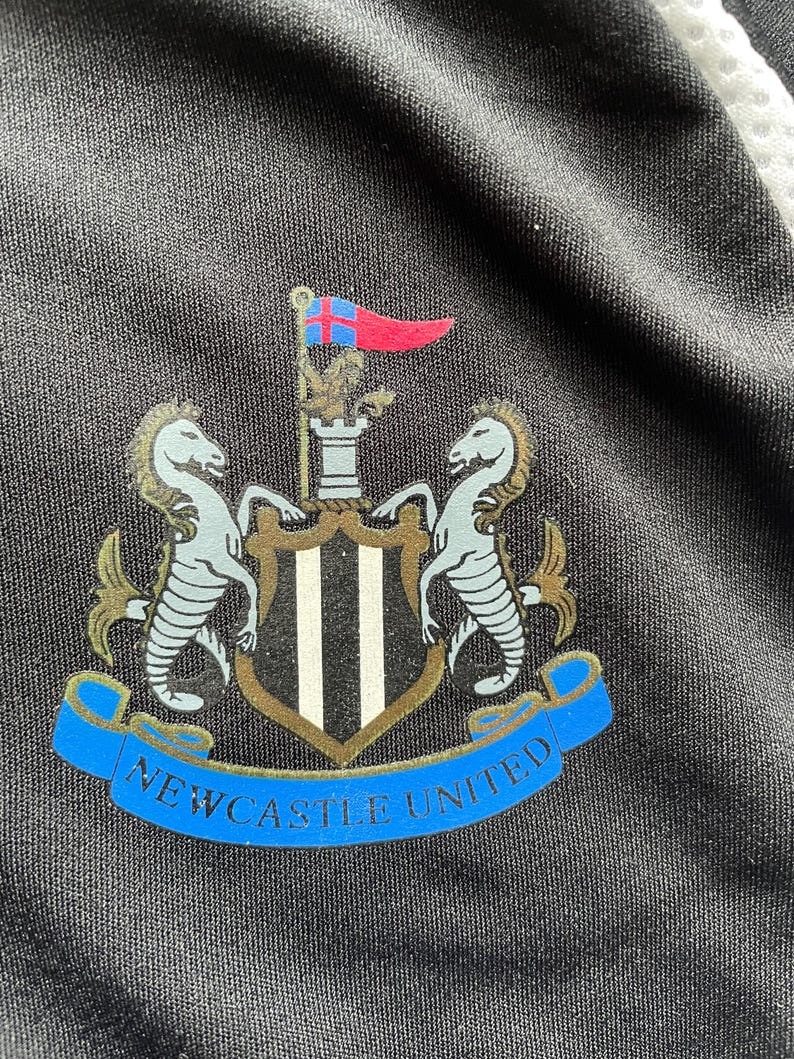 Pu&ograve; includere: Primo piano di un tessuto nero con lo stemma del Newcastle United. Lo stemma presenta due cavallucci marini, uno scudo a strisce bianche e nere e uno stendardo blu con la scritta "Newcastle United". Una bandiera rossa e blu in alto.