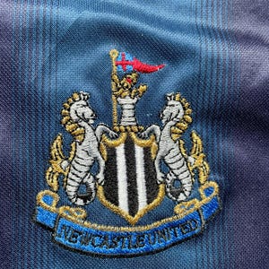 May include: Embroidered Newcastle United crest on a blue and black striped fabric. The crest features two seahorses, a shield with black and white stripes, and a banner that reads "NEWCASTLE UNITED". A red and blue flag is at the top.