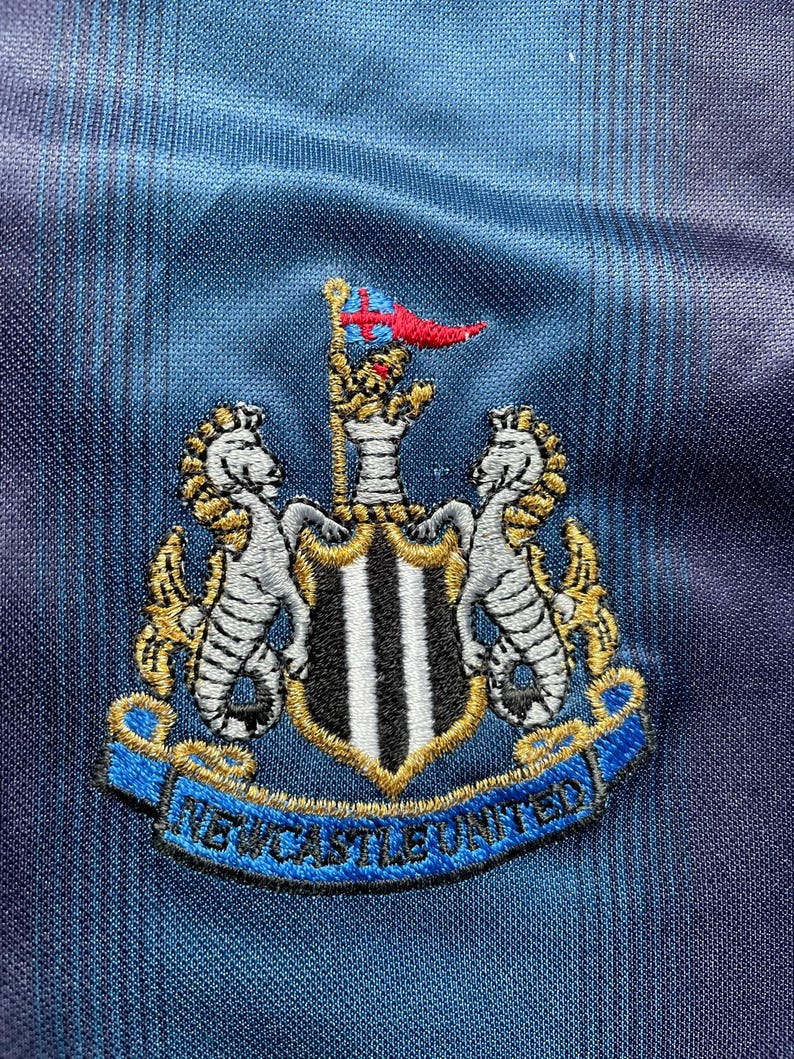 May include: Embroidered Newcastle United crest on a blue and black striped fabric. The crest features two seahorses, a shield with black and white stripes, and a banner that reads "NEWCASTLE UNITED". A red and blue flag is at the top.