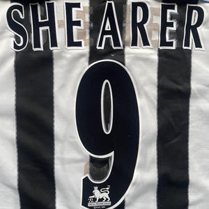 May include: A white and black striped soccer jersey with the name "SHEARER" and the number "9" on the back. The jersey also features a Premier League logo.