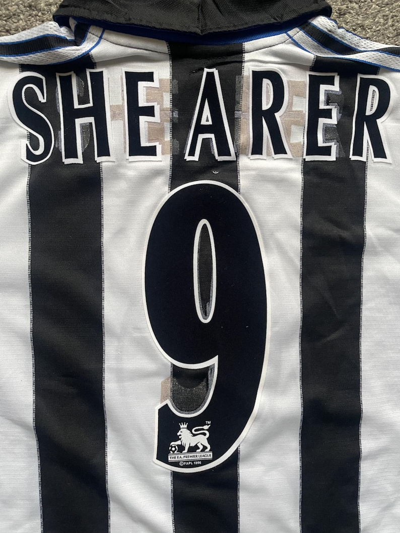 May include: A white and black striped soccer jersey with the name "SHEARER" and the number "9" on the back. The jersey also features a Premier League logo.