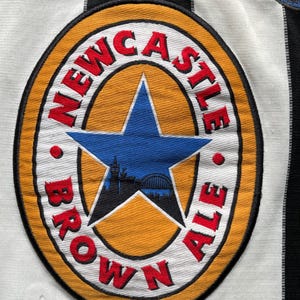 Puede incluir: Una camiseta de f&uacute;tbol a rayas blancas y negras con un logotipo de Newcastle Brown Ale en la parte delantera. El logotipo es un c&iacute;rculo amarillo con una estrella roja y las palabras "Newcastle Brown Ale" alrededor.