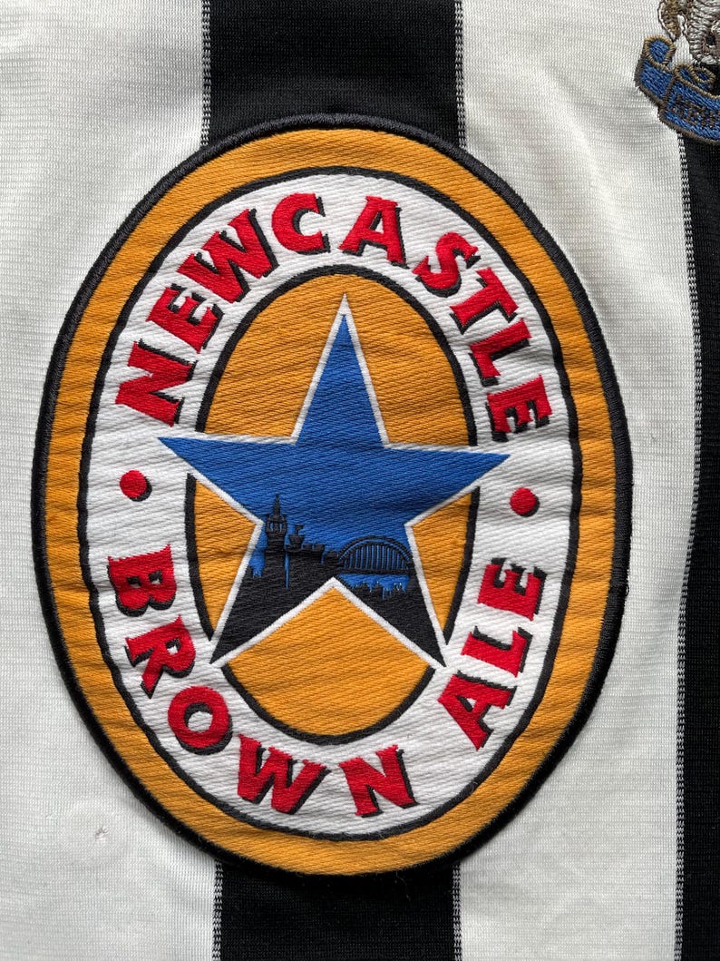 Puede incluir: Una camiseta de f&uacute;tbol a rayas blancas y negras con un logotipo de Newcastle Brown Ale en la parte delantera. El logotipo es un c&iacute;rculo amarillo con una estrella roja y las palabras "Newcastle Brown Ale" alrededor.