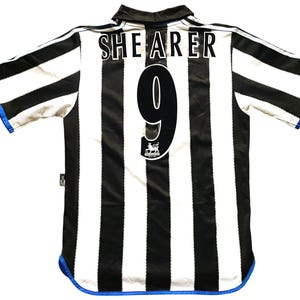 May include: A black and white striped soccer jersey with the name "SHEARER" and the number "9" on the back. The jersey has a blue trim on the sleeves and bottom hem. A Premier League logo is visible.