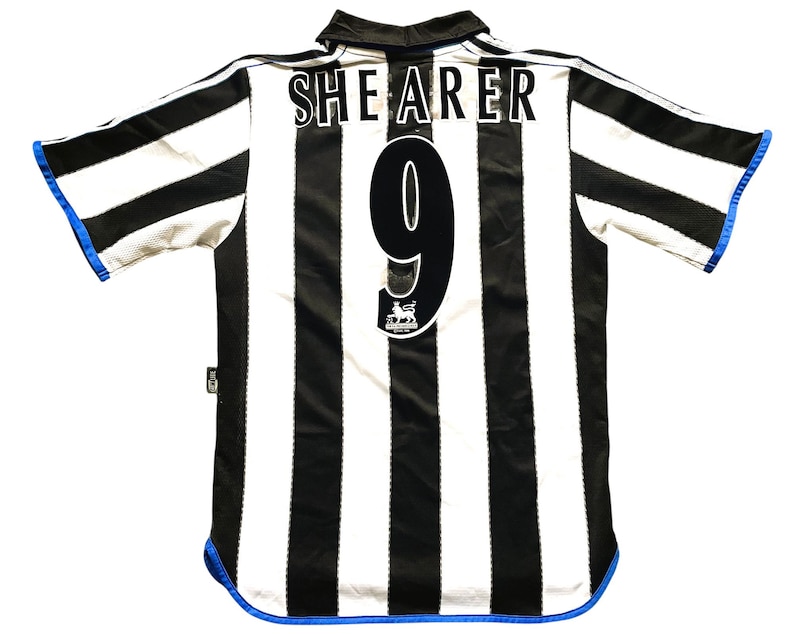 May include: A black and white striped soccer jersey with the name "SHEARER" and the number "9" on the back. The jersey has a blue trim on the sleeves and bottom hem. A Premier League logo is visible.