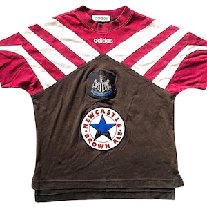Op de afbeelding: Een vintage Adidas Newcastle United voetbalshirt. Het shirt heeft bordeauxrode en witte strepen op de schouders en mouwen, met een bruin voorpaneel. Het Newcastle Brown Ale logo en het teamembleem zijn duidelijk zichtbaar.