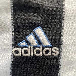 May include: Close-up of a black and white striped fabric with the Adidas logo embroidered in white with light blue accents. The logo features three angled stripes above the brand name.