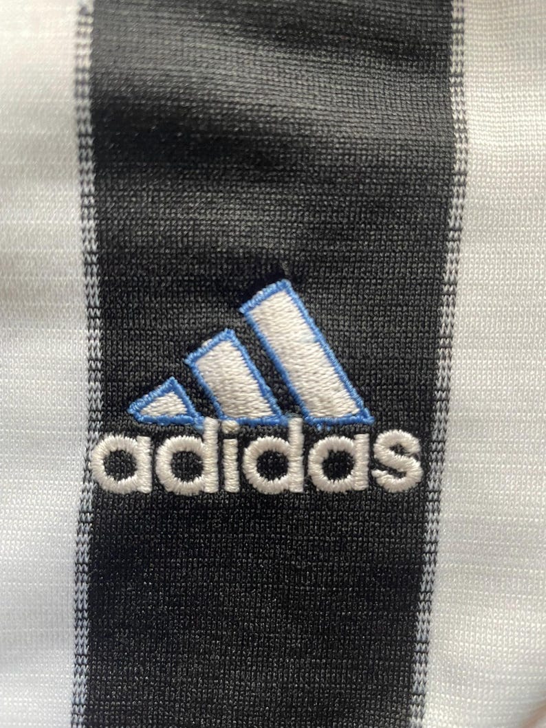 May include: Close-up of a black and white striped fabric with the Adidas logo embroidered in white with light blue accents. The logo features three angled stripes above the brand name.