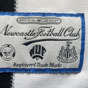 May include: A rectangular label with blue trim, featuring the text "Newcastle Football Club" in elegant script. The label includes several small emblems and the words "Official Garment" and "Registered Trade Marks".