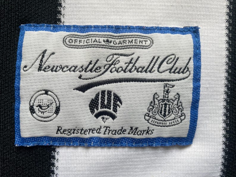 May include: A rectangular label with blue trim, featuring the text "Newcastle Football Club" in elegant script. The label includes several small emblems and the words "Official Garment" and "Registered Trade Marks".