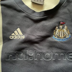 Puede incluir: Una camiseta de f&uacute;tbol a rayas blancas y negras con el logotipo de Adidas, el escudo del Newcastle United y la palabra "nufc.home" bordada en la parte delantera.