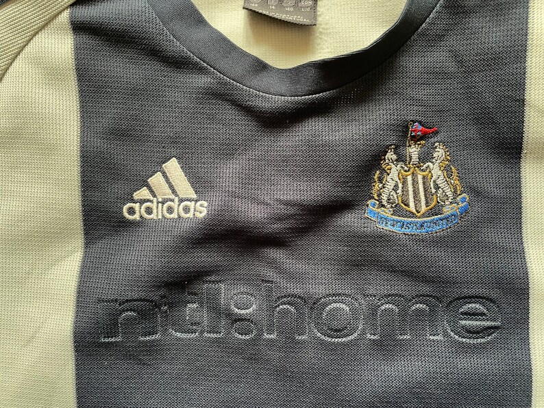 Puede incluir: Una camiseta de f&uacute;tbol a rayas blancas y negras con el logotipo de Adidas, el escudo del Newcastle United y la palabra "nufc.home" bordada en la parte delantera.