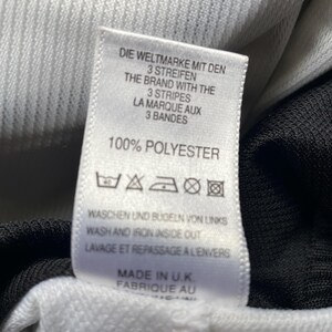 May include: White tag with black text that reads "Die Weltmarke mit den 3 Streifen", "The Brand with the 3 Stripes", "La Marque aux 3 Bandes", "100% Polyester". The tag also includes washing instructions and "Made in U.K. Fabrique au".