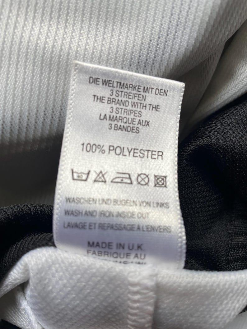 May include: White tag with black text that reads "Die Weltmarke mit den 3 Streifen", "The Brand with the 3 Stripes", "La Marque aux 3 Bandes", "100% Polyester". The tag also includes washing instructions and "Made in U.K. Fabrique au".