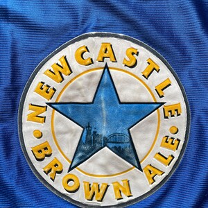 K&ouml;nnte beinhalten: Ein blauer und gelber runder Patch mit dem Text "Newcastle Brown Ale" um einen blauen f&uuml;nfzackigen Stern.