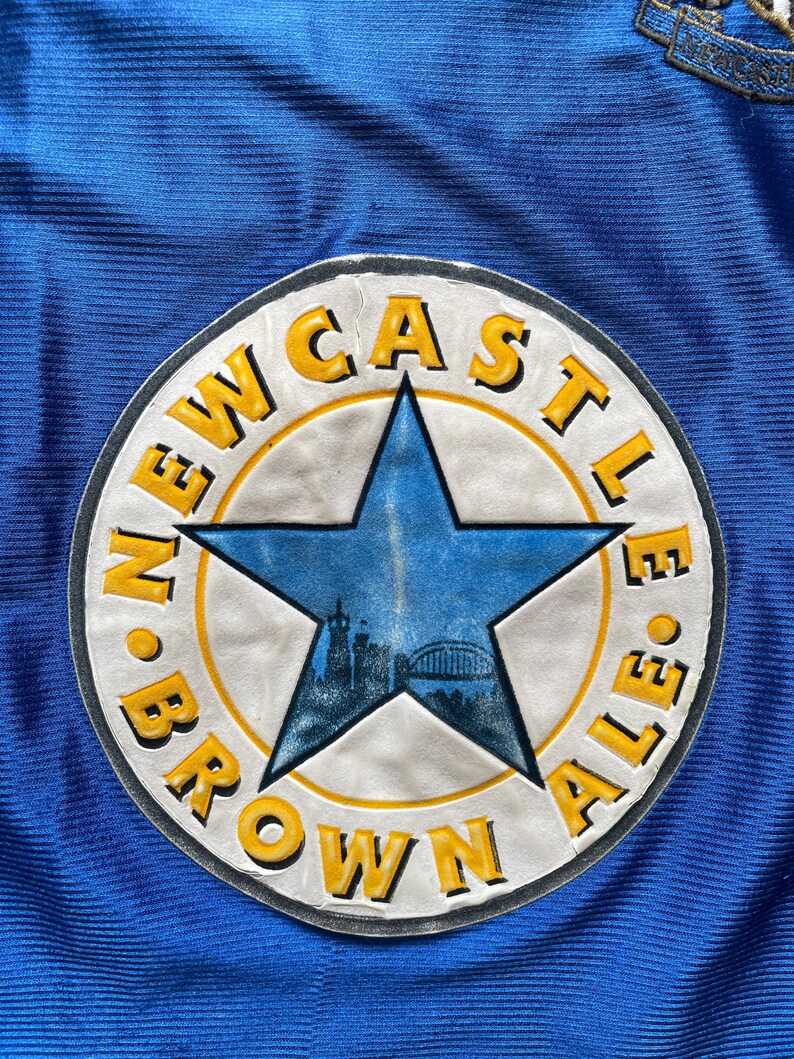 K&ouml;nnte beinhalten: Ein blauer und gelber runder Patch mit dem Text "Newcastle Brown Ale" um einen blauen f&uuml;nfzackigen Stern.