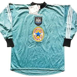 May include: A teal long-sleeve soccer jersey with white stripes on the sleeves and black cuffs. The jersey features the Newcastle United crest and a Newcastle Brown Ale logo. An Adidas tag is attached.