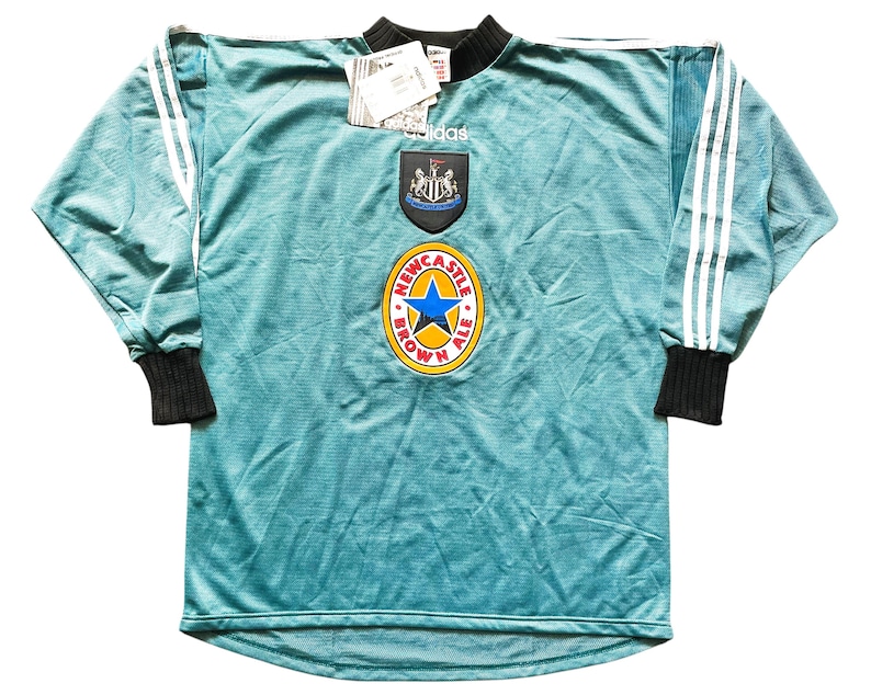 May include: A teal long-sleeve soccer jersey with white stripes on the sleeves and black cuffs. The jersey features the Newcastle United crest and a Newcastle Brown Ale logo. An Adidas tag is attached.