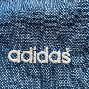 May include: Close-up of blue fabric with the white embroidered Adidas logo. The fabric has a textured appearance with a subtle pattern. The Adidas logo is in a classic, clean font.