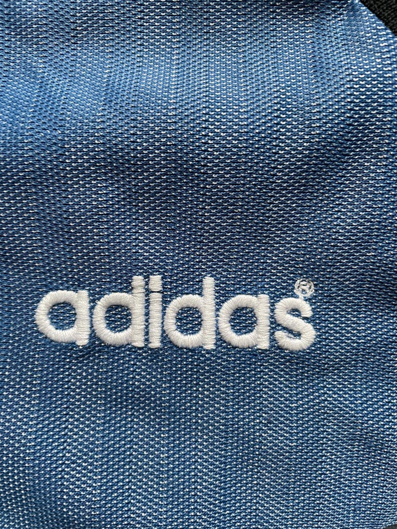 May include: Close-up of blue fabric with the white embroidered Adidas logo. The fabric has a textured appearance with a subtle pattern. The Adidas logo is in a classic, clean font.