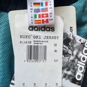 K&ouml;nnte beinhalten: Nahaufnahme eines Adidas-Etiketts, das an einem t&uuml;rkis-schwarzen Trikot befestigt ist. Das Etikett zeigt das Adidas-Logo, Flaggen und Text, darunter "NUFC GK1 JERSEY" und einen Barcode. Das Trikot ist teilweise sichtbar.
