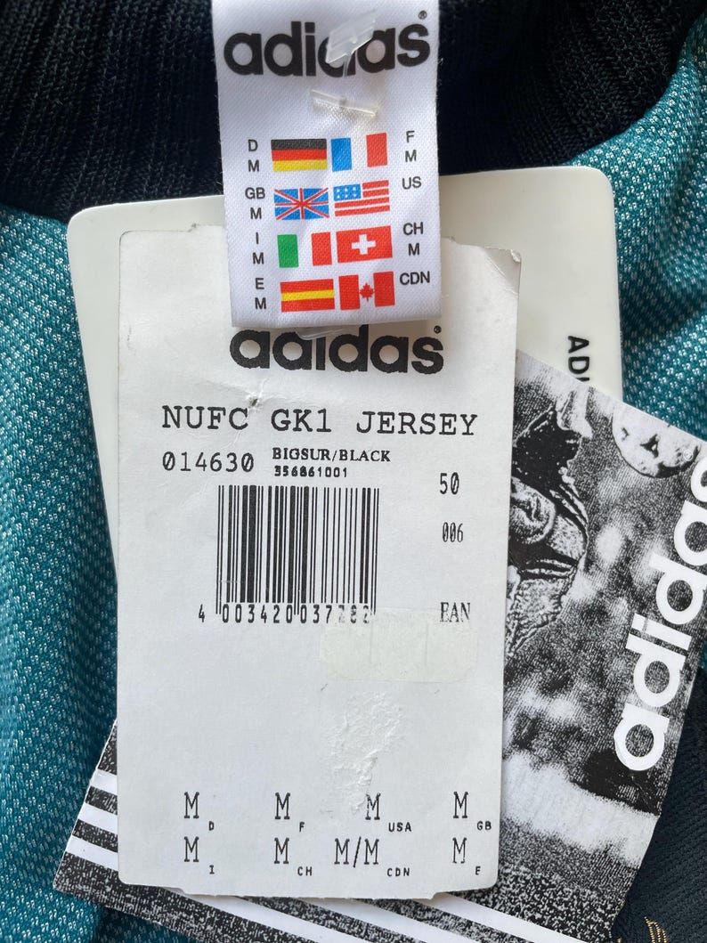 K&ouml;nnte beinhalten: Nahaufnahme eines Adidas-Etiketts, das an einem t&uuml;rkis-schwarzen Trikot befestigt ist. Das Etikett zeigt das Adidas-Logo, Flaggen und Text, darunter "NUFC GK1 JERSEY" und einen Barcode. Das Trikot ist teilweise sichtbar.