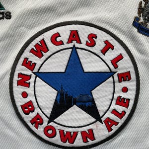May include: A white patch with a black circle and a red and blue star. The circle has the text "NEWCASTLE BROWN ALE" around the edge.