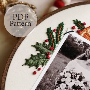 May include: A close-up of a white fabric with green and red embroidery of holly leaves and berries. A black and white photo is partially visible in the background.