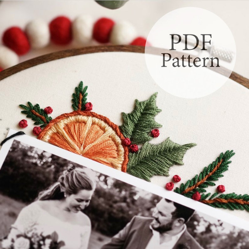 May include: Embroidered orange slice with holly and berry sprigs on a white fabric background. The fabric is stretched over a wooden embroidery hoop. The text "PDF Pattern" is written in a white circle on a white background.