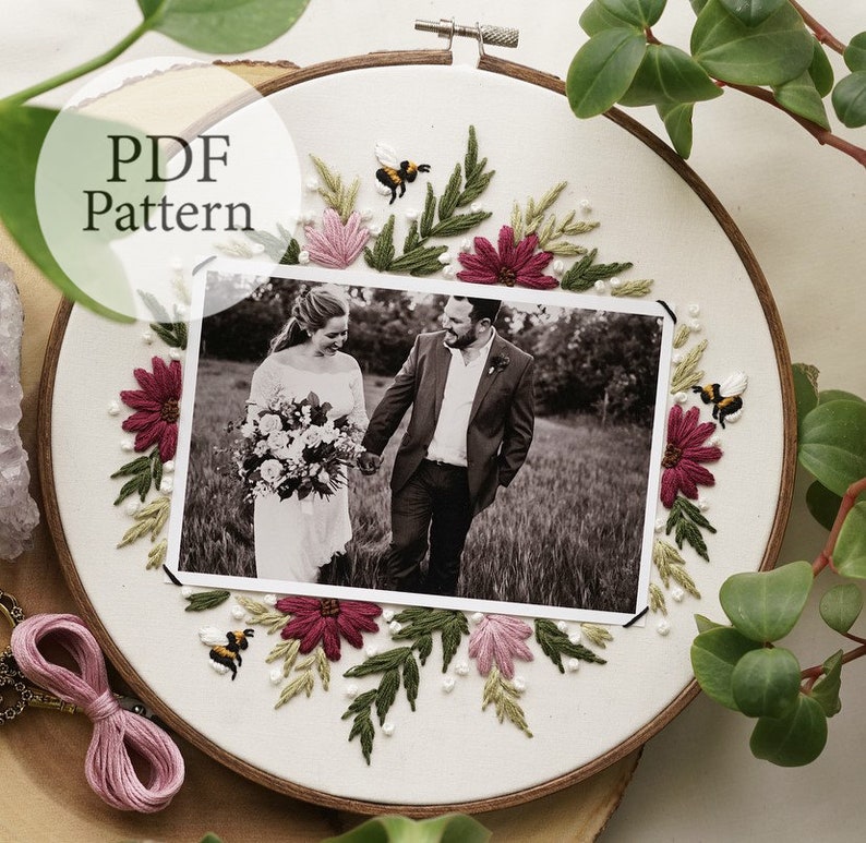May include: A white embroidery hoop with a black and white photo of a couple in the center. The photo is surrounded by embroidered flowers, leaves, and bees in shades of pink, green, and yellow.