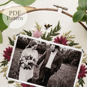 May include: A white embroidery hoop with a black and white photo of a couple in a field. The photo is surrounded by embroidered flowers and leaves in shades of pink, green, and burgundy. There is a small embroidered bee in the upper right corner of the hoop. The text "PDF Pattern" is written in black on a white circle in the upper left corner of the hoop.