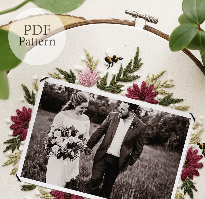 May include: A white embroidery hoop with a black and white photo of a couple in a field. The photo is surrounded by embroidered flowers and leaves in shades of pink, green, and burgundy. There is a small embroidered bee in the upper right corner of the hoop. The text "PDF Pattern" is written in black on a white circle in the upper left corner of the hoop.