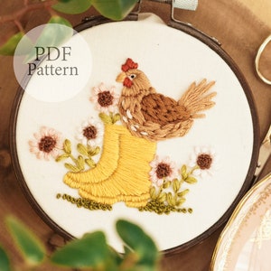 May include: Embroidered hoop art featuring a brown chicken perched on a pair of yellow wellies with white and brown flowers surrounding them. The hoop is made of wood and has a white fabric background. The text "PDF Pattern" is written in black at the top of the hoop.