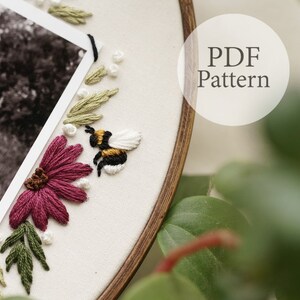 May include: Embroidered hoop art with a bee, flowers, and leaves. The hoop is made of wood and has a white fabric background. The bee is yellow and black with white wings. The flowers are pink and the leaves are green. The text "PDF Pattern" is visible in the upper right corner of the image.