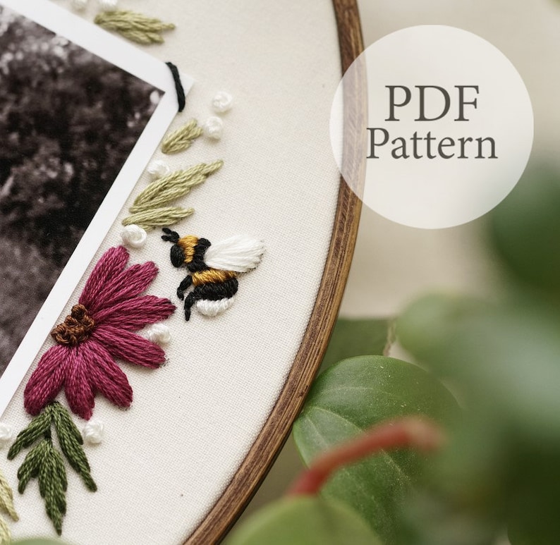May include: Embroidered hoop art with a bee, flowers, and leaves. The hoop is made of wood and has a white fabric background. The bee is yellow and black with white wings. The flowers are pink and the leaves are green. The text "PDF Pattern" is visible in the upper right corner of the image.