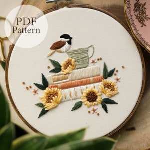 Op de afbeelding: Borduurwerk op een borduurring met een stapel boeken met een kleine vogel die op een theekopje zit. De boeken zijn versierd met zonnebloemen en bladeren. De achtergrond is een witte stof.