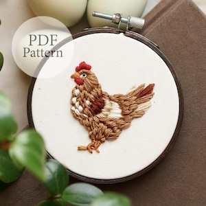 Könnte beinhalten: Gestickter brauner Hahn mit weißen und roten Akzenten auf einem weißen Stoff in einem hölzernen Stickrahmen. Der Rahmen befindet sich auf einer braunen Oberfläche. Der Text "PDF Pattern" befindet sich in einem weißen Kreis über dem Rahmen.