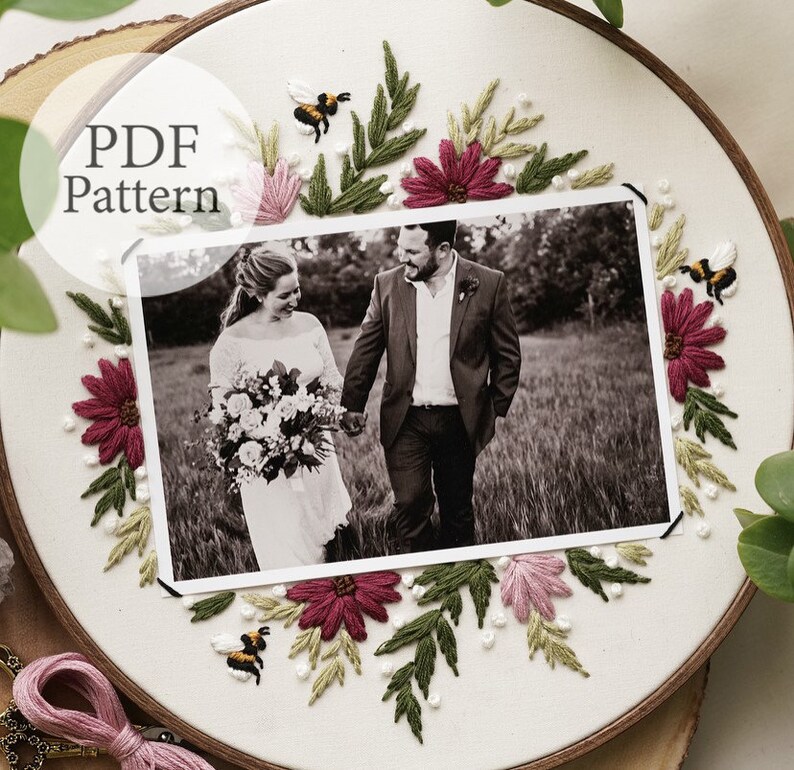 May include: A white embroidery hoop with a black and white photo of a couple in the center. The photo is surrounded by embroidered flowers, leaves, and bees in shades of pink, green, and yellow.