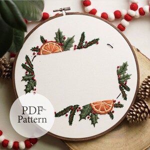 May include: A white embroidery hoop with a Christmas-themed design. The design features green holly leaves, red berries, and orange citrus slices. The text "PDF Pattern" is visible in the lower left corner of the image.