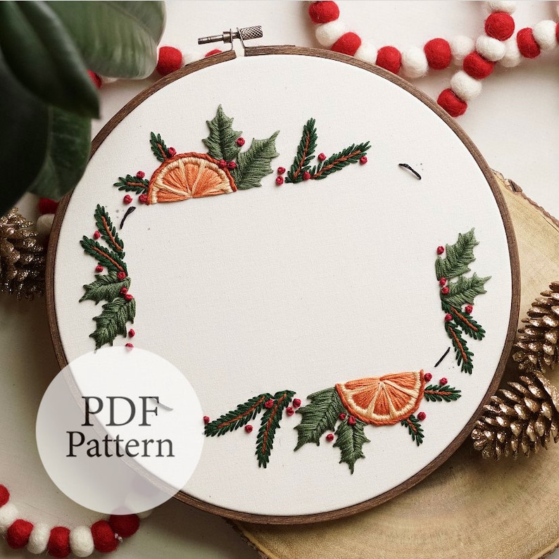 May include: A white embroidery hoop with a Christmas-themed design. The design features green holly leaves, red berries, and orange citrus slices. The text "PDF Pattern" is visible in the lower left corner of the image.