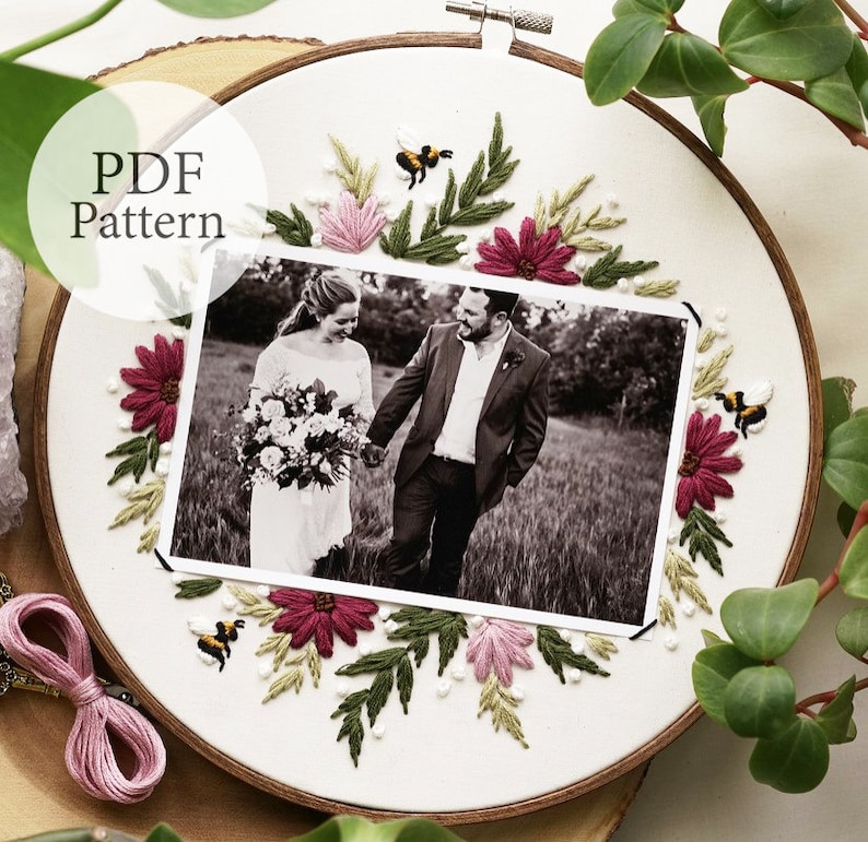 May include: A white embroidery hoop with a black and white photo of a couple in the center. The photo is surrounded by embroidered flowers in shades of pink and red, green leaves, and small yellow bees.