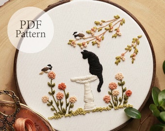 May include: Embroidered hoop art featuring a black cat sitting on a white mushroom with two small birds perched on branches. The background is a white fabric with green and peach floral embroidery.