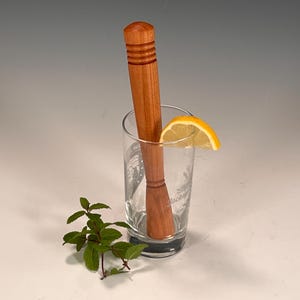 May include: A wooden cocktail muddler with a textured top, resting in a clear glass with a lemon wedge and a sprig of mint.